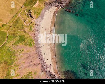 Vista aerea dall'alto di una spiaggia sabbiosa curvata lungo la costa turchese, scogliere rossastre, promontorio erboso e onde gentili che si infrangono sulla riva in estate Foto Stock