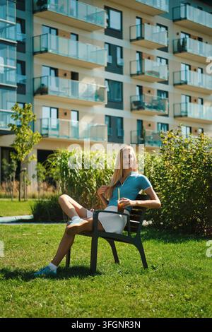 Donna si rilassa su una sedia nel giardino di una zona residenziale indossando una camicia blu e una gonna bianca. Godeva una bevanda fredda da un bicchiere da asporto in una giornata soleggiata Foto Stock