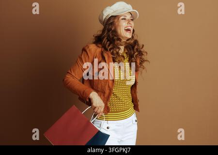Una donna sorridente, vestita con moda autunnale con una giacca e un berretto arancione, posa su uno sfondo marrone caldo. Tiene felicemente borse della spesa, embo Foto Stock