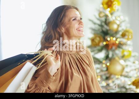 Donna raggiante in maglione a collo alto di raso dorato che brilla, porta borse della spesa. Un albero di Natale sfocato con ornamenti dorati e luci creates una luce brillante Foto Stock
