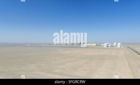 Veduta aerea di un moderno stabilimento per la lavorazione dei materiali in silicio con laboratori bianchi, torri ed edifici residenziali isolati nel piatto e arido deserto del Gobi sotto un cielo blu chiaro, con una strada rettilinea che conduce all'orizzonte, provincia di Gansu, Cina. Foto Stock