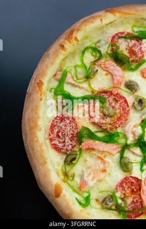 Una pizza rotonda poggia su una superficie nera. I condimenti includono fette di pomodoro, olive ed erbe verdi. La crosta è dorata e leggermente croccante. Foto Stock