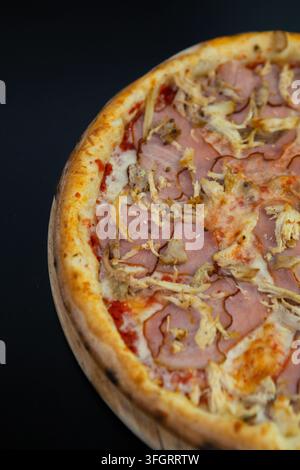 Una pizza rotonda è posata su un piatto di legno. La pizza è ricoperta da fette di prosciutto e pezzi di pollo su formaggio fuso e salsa di pomodoro. Questo piatto è re Foto Stock