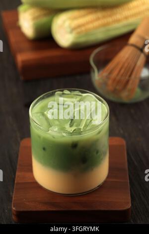 Bevanda ghiacciata stratificata al tè verde matcha con latte di mais in un bicchiere trasparente, con un pannocchia di mais sullo sfondo Foto Stock