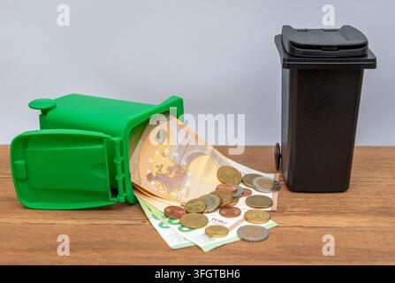 Banconote e monete in euro fuoriescono da bidoni della spazzatura verdi e neri rovesciati su una superficie di legno. Foto Stock