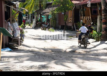 Un motociclista percorre una strada sterrata sabbiosa fiancheggiata da negozi rustici in legno e edifici con tetti di paglia in un tranquillo villaggio tropicale, probabilmente in Thailandia Foto Stock