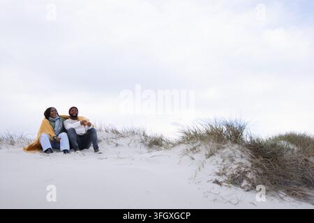 Coppia seduta su duna di sabbia in spiaggia avvolta in una coperta gialla con berretto bordeaux, spazio libero Foto Stock
