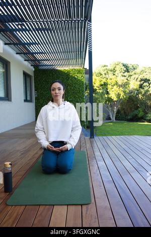 Donna adulta in ginocchio su un tappeto da yoga verde sul ponte con felpa con cappuccio e pantaloni blu-verdi, bottiglia nelle vicinanze Foto Stock