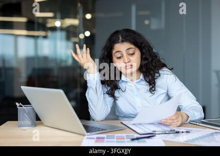 Donna d'affari stressata che lavora su laptop e documenti in ufficio mentre si sente sopraffatta dai compiti di analisi finanziaria. Una manager professionista che legge documenti cartacei alla sua scrivania. Foto Stock