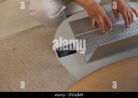 Donna anziana afroamericana che digita su un laptop argentato a casa, carta di credito nelle vicinanze Foto Stock