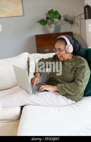 Donna afroamericana anziana seduta su un divano color crema a casa, indossa un auricolare rosa e digita su un laptop Foto Stock