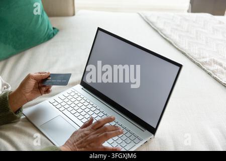 Donna anziana afroamericana che tiene una carta di credito, digita su un laptop a letto e indossa un maglione verde Foto Stock