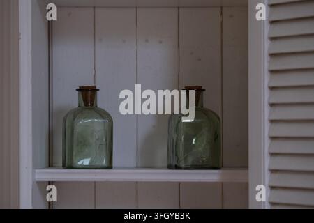 Due bottiglie di vetro verde vintage esposte su uno scaffale di legno bianco, creando una semplice natura morta rustica. Decorazione domestica minimale e stile interno con r Foto Stock