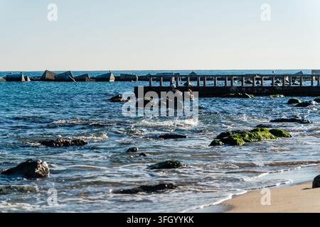 Vista della costa rocciosa e del molo in cemento sulla spiaggia con pietre ricoperte di muschio e onde calme del mar Mediterraneo a Barcellona, Catalogna, Spagna. Foto Stock