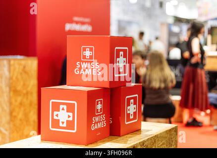 Colonia, Germania - 26 agosto 2026: Cubi rossi impilati con il marchio Swiss Games su una piattaforma di legno con figure sfocate di partecipanti sullo sfondo Foto Stock