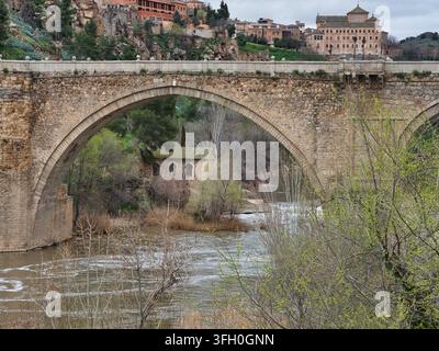 Storico ponte di pietra sul fiume Tago - Il maestoso Puente de San Martín, un ponte ad arco medievale, si estende sul fiume Tago con un edificio in pietra visibile Foto Stock