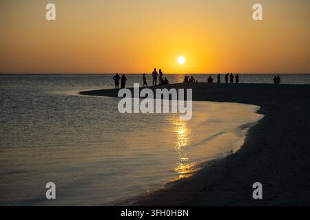 Una fotografia paesaggistica ampia che cattura un gruppo di persone in controluce davanti a un tramonto vivace sul mare. Gli individui e i piccoli gruppi sono riuniti Foto Stock