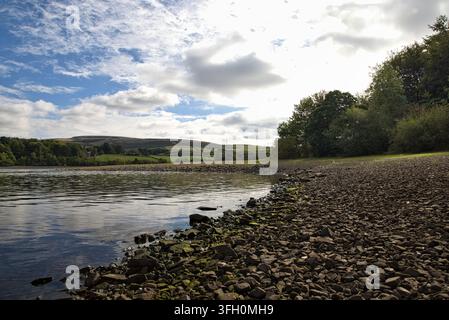 La luce del sole che filtra attraverso i cieli nuvolosi sopra la riva ghiaiosa e le acque calme del lago Hollingworth, Rochdale, UK. Foto Stock