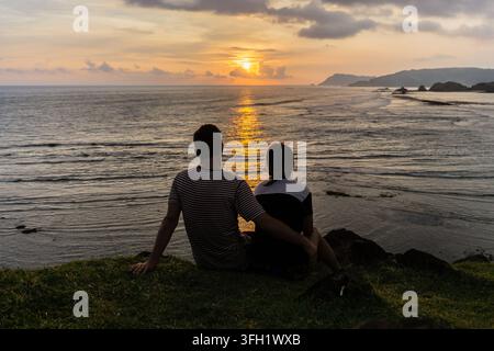 Due giovani innamorati si siedono in cima a una scogliera per guardare il tramonto sull'oceano a Lombok, Indonesia Foto Stock