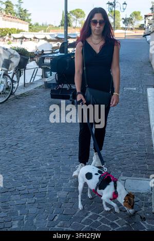 Una bella donna con lunghi capelli scuri, vestita di nero, passeggia col suo cane Jack Russell nel centro storico di una piccola città Foto Stock