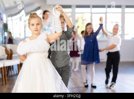 Entusiasti preadolescenti, ragazzi e ragazze in abbigliamento da festa che eseguono eleganti valzer a coppie nella soleggiata sala della scuola di ballo con teac sorridente Foto Stock
