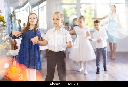 Preadolescenti entusiasti, ragazze e ragazzi in abiti eleganti che praticano passi di danza di coppia in uno studio adornato di decorazioni natalizie, sotto Gu Foto Stock