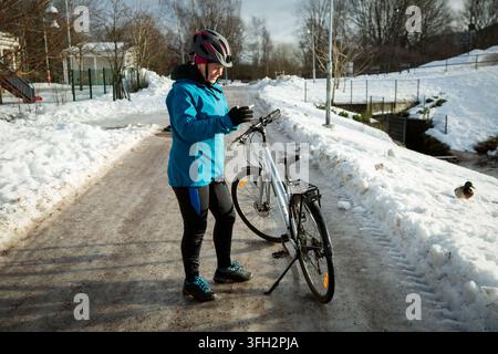 Una donna attiva di mezza età controlla il suo smartphone accanto a una bicicletta su una strada nevosa di un parco invernale, pianificando il suo tragitto in bicicletta. Foto Stock