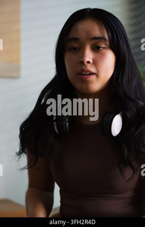 Donna afroamericana che parla, indossa cuffie over-ear al collo in una stanza con pannelli di legno Foto Stock