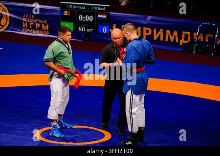 Lottatori che si affrontano in una gara competitiva con l'arbitro su un tappeto blu durante il torneo internazionale, World Nomad Games 2026, Kirghizistan. Foto Stock