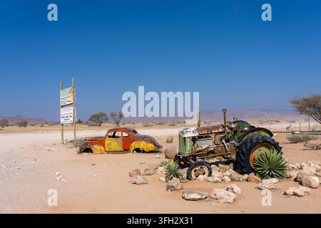 Trattore vintage arrugginito e auto abbandonata accanto ai segnali stradali in un paesaggio rurale arido della regione di Kunene, Namibia, con montagne sullo sfondo. Foto Stock