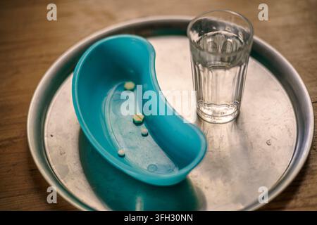 Disposizione da tavolo di varie pillole in una ciotola renale turchese accanto a un bicchiere d'acqua su un vassoio medico in metallo. Foto Stock