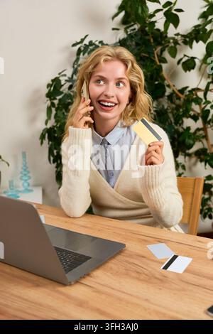 Una donna bionda si gode lo shopping online alla sua scrivania, tenendo una carta di credito e al telefono. Foto Stock