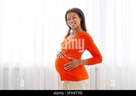 Donna nera incinta sorridente in piedi vicino alla finestra a casa, toccandosi delicatamente la pancia Foto Stock