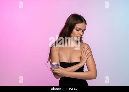 Giovane donna che applica una crema per il corpo, nutre la pelle con una crema idratante per la bellezza e il benessere, su uno sfondo sfumato Foto Stock