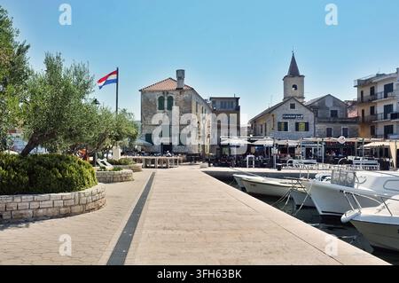 Pirovac, Croazia, 16 agosto 2026 Promenada tranquilla sul lungomare accanto a barche ormeggiate, antichi edifici in pietra, campanile della chiesa, terrazze di ristoranti e bandiera su s Foto Stock