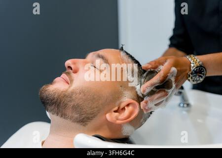Prima di un uomo barbuto con gli occhi chiusi che riceve un rilassante lavaggio dei capelli con shampoo da un barbiere a un lavandino bianco in un salone di barbiere moderno. Foto Stock