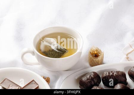 Prima di sacchetti di tè alle erbe in una tazza bianca accanto a una candela a forma di miele, caramelle al cioccolato fondente e una noce su un tessuto bianco. La luce naturale calda crea un Foto Stock