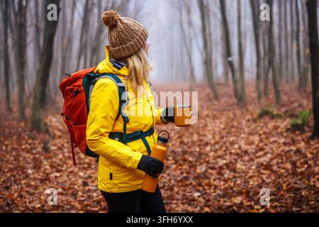 Escursionista in giacca gialla con zaino che tiene un thermos e una tazza da viaggio in una foresta autunnale. Rinfresco durante il trekking in condizioni di freddo e nebbia Foto Stock