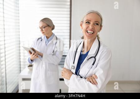 Donna medico di mezza età sicura di sé che ride in piedi nell'ufficio medico della clinica Foto Stock