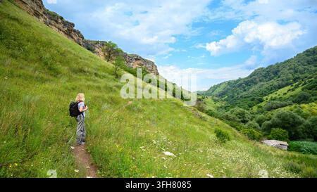 Una donna bionda con una maglietta azzurra, pantaloni grigi e uno zaino nero si trova su un sentiero stretto di terra su una collina erbosa ripida. Sta guardando Foto Stock