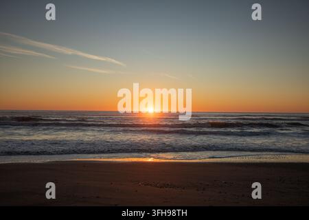 Sole dorato che tramonta su onde oceaniche ondulate con una spiaggia sabbiosa e un cielo serale caldo Foto Stock