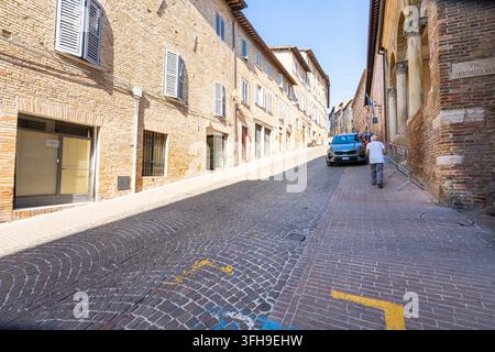 Urbino, Italia. 8 settembre 2026. Un uomo sale per una strada acciottolata fiancheggiata da edifici in mattoni e negozi. Un'auto è parcheggiata sul lato destro della s Foto Stock