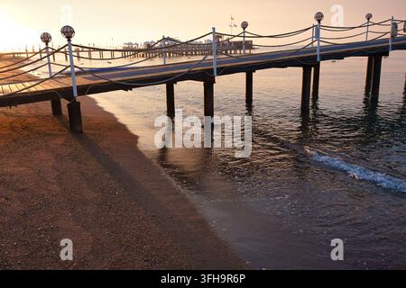 Una vista ad angolo alto di un molo di legno con ringhiere di corda e lampade a globo che si estende da una spiaggia di sabbia bagnata verso dolci onde oceaniche all'alba. Oro morbido Foto Stock