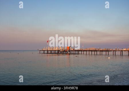Una vasta vista panoramica di un lungo molo di legno con lettini, ombrelloni e una bandiera turca che si estende nelle acque calme dell'oceano all'alba. Il primo mattino Foto Stock