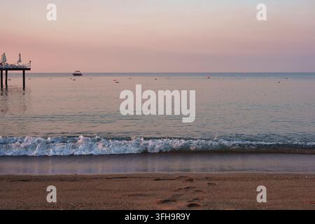 Un paesaggio costiero calmo con un'onda schiumosa e gentile che lambisce una riva sabbiosa durante un'alba tranquilla. Un bordo del molo in legno è visibile sulla l Foto Stock