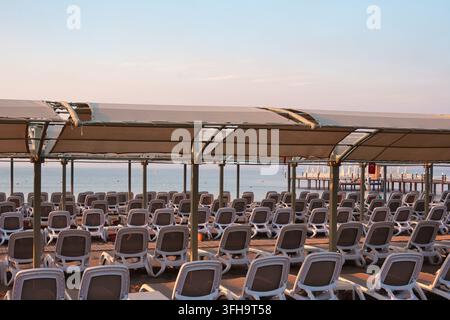 Una vista ampia da dietro file di sdraio bianchi vuoti sotto una struttura ombreggiata sulla spiaggia, rivolti verso l'oceano calmo all'alba. Luce delicata del mattino Foto Stock