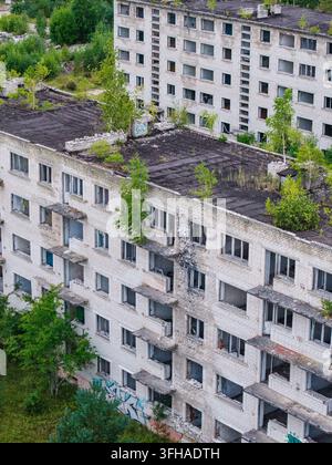 Veduta aerea della città militare sovietica abbandonata di Irbene, con edifici residenziali in rovina su cui crescono alberi sui tetti e graffiti sui muri a I Foto Stock