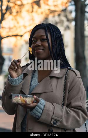 Donna nera felice che mangia un'insalata di frutta da asporto su una strada cittadina in autunno Foto Stock