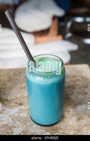 RIGA, LETTONIA. 25 agosto 2026. Un latte matcha alla spirulina blu in un bicchiere trasparente con una cannuccia riutilizzabile in metallo, servito su un tavolo di marmo in un caffè moderno. Foto Stock