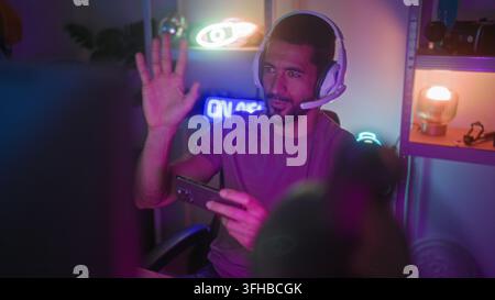 Uomo con auricolare e microfono tiene uno smartphone con entrambe le mani, gesticola con la mano sinistra e sorride seduto in uno studio di gaming al neon; eccitazione Foto Stock